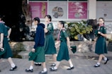 A group of young girls wearing green uniforms, likely school or scout attire, are walking on a tiled pathway. They are in an outdoor area with a backdrop featuring posters and lush green plants. One girl is wearing a dark blue hoodie while others are in short-sleeved shirts, holding hats in their hands.