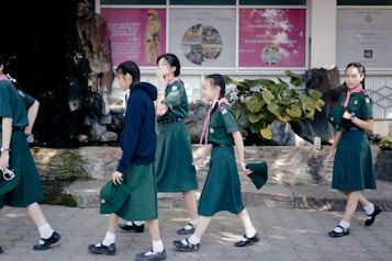 A group of young girls wearing green uniforms, likely school or scout attire, are walking on a tiled pathway. They are in an outdoor area with a backdrop featuring posters and lush green plants. One girl is wearing a dark blue hoodie while others are in short-sleeved shirts, holding hats in their hands.