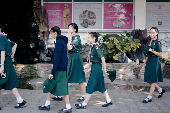 A group of young girls wearing green uniforms, likely school or scout attire, are walking on a tiled pathway. They are in an outdoor area with a backdrop featuring posters and lush green plants. One girl is wearing a dark blue hoodie while others are in short-sleeved shirts, holding hats in their hands.