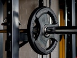 A close-up view of a barbell with a 10 kg weight plate attached. The barbell is positioned on a rack, and the background shows part of a gym environment with metallic and brick textures.