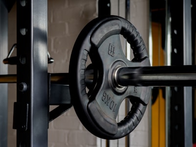 A close-up view of a barbell with a 10 kg weight plate attached. The barbell is positioned on a rack, and the background shows part of a gym environment with metallic and brick textures.