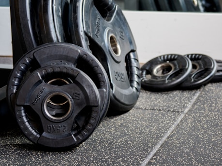 A collection of black weight plates of varying sizes rests on a gym floor with a speckled texture. The weight plates are stacked and positioned against a reflective surface. The central weight plate prominently displays the weight measurement and brand.