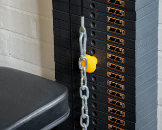 A close-up view of a weight stack used in gym equipment, with labels indicating different weights in pounds and kilograms. A yellow pin is inserted into the stack to select the desired weight, and a metal chain is connected to it. The background includes a white brick wall and a padded black seat.