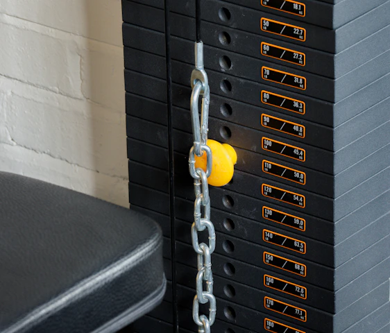 A close-up view of a weight stack used in gym equipment, with labels indicating different weights in pounds and kilograms. A yellow pin is inserted into the stack to select the desired weight, and a metal chain is connected to it. The background includes a white brick wall and a padded black seat.