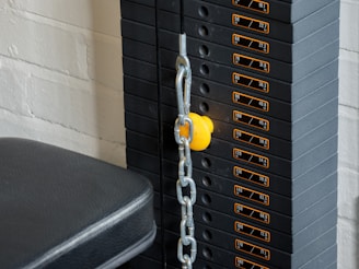 A close-up view of a weight stack used in gym equipment, with labels indicating different weights in pounds and kilograms. A yellow pin is inserted into the stack to select the desired weight, and a metal chain is connected to it. The background includes a white brick wall and a padded black seat.
