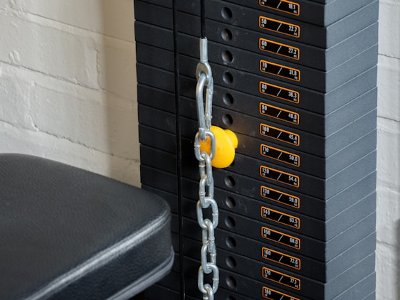 A close-up view of a weight stack used in gym equipment, with labels indicating different weights in pounds and kilograms. A yellow pin is inserted into the stack to select the desired weight, and a metal chain is connected to it. The background includes a white brick wall and a padded black seat.