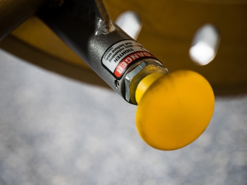 A close-up image of an industrial valve or handle with a bright yellow knob. The metal component features a warning label indicating danger and instructions, likely part of a machinery or equipment.