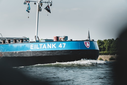 A large blue and black ship, labeled 'EILTANK 47,' is moving along a body of water. Several flags are flying on poles attached to the ship. The shoreline in the background shows greenery and a distant church spire.
