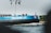 A large blue and black ship, labeled 'EILTANK 47,' is moving along a body of water. Several flags are flying on poles attached to the ship. The shoreline in the background shows greenery and a distant church spire.