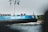 A large blue and black ship, labeled 'EILTANK 47,' is moving along a body of water. Several flags are flying on poles attached to the ship. The shoreline in the background shows greenery and a distant church spire.