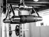 A close-up of a metal exercise machine handle with a textured grip, designed for fitness routines. The background features elements of a gym, including weight equipment, blurred out to emphasize the handle.
