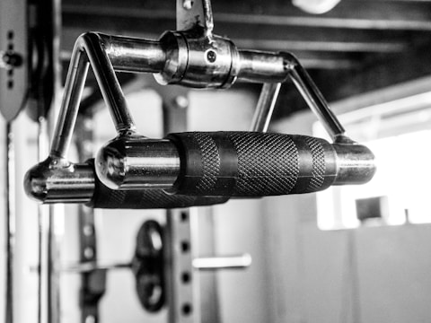 A close-up of a metal exercise machine handle with a textured grip, designed for fitness routines. The background features elements of a gym, including weight equipment, blurred out to emphasize the handle.
