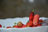 A white ceramic bowl contains a collection of ripe red and pink mulberries with some green stems visible. The fruit is piled casually, creating a vibrant display of natural textures and colors. Ice cubes are present in the bowl, adding a fresh and cool element.