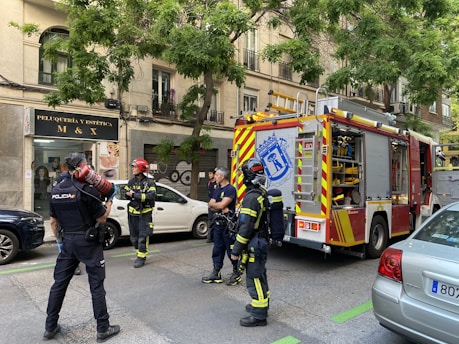 Firefighters and a police officer stand on a city street near a fire truck. The setting is urban, with a building that has a hair salon sign in the background. The firefighters are in their gear, and the fire truck is equipped with emergency tools. A police officer holds a fire extinguisher, indicating a possible emergency response scenario.