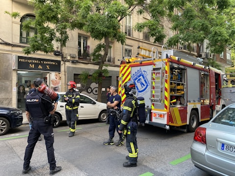 Firefighters and a police officer stand on a city street near a fire truck. The setting is urban, with a building that has a hair salon sign in the background. The firefighters are in their gear, and the fire truck is equipped with emergency tools. A police officer holds a fire extinguisher, indicating a possible emergency response scenario.