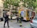 Firefighters and a police officer stand on a city street near a fire truck. The setting is urban, with a building that has a hair salon sign in the background. The firefighters are in their gear, and the fire truck is equipped with emergency tools. A police officer holds a fire extinguisher, indicating a possible emergency response scenario.
