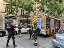 Firefighters and a police officer stand on a city street near a fire truck. The setting is urban, with a building that has a hair salon sign in the background. The firefighters are in their gear, and the fire truck is equipped with emergency tools. A police officer holds a fire extinguisher, indicating a possible emergency response scenario.