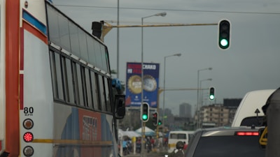 A busy urban street with several vehicles, including a large bus and cars, waiting at a traffic light showing green. Traffic lights are visible over the road, and a billboard can be seen in the background with buildings beyond.