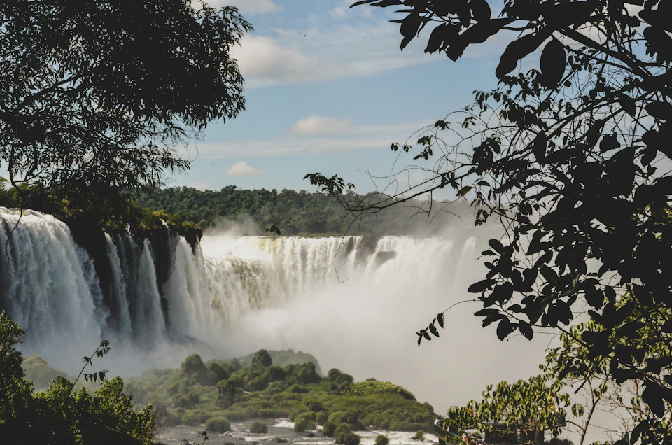 The image depicts a majestic waterfall cascading down into a pool below, surrounded by lush green trees and bushes. The waterfall is situated on the left side of the frame, with its powerful flow creating a misty spray that adds to the overall atmosphere. On the right side, there are several trees and bushes, their leaves forming a natural canopy over the scene. The sky above is clear blue, providing a beautiful backdrop for this picturesque landscape.
