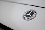 The image features a close-up of a Mercedes-Benz logo on a smooth, light-colored surface. The logo is in the shape of a three-pointed star within a circle, with the brand name inscribed around it.