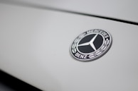 The image features a close-up of a Mercedes-Benz logo on a smooth, light-colored surface. The logo is in the shape of a three-pointed star within a circle, with the brand name inscribed around it.