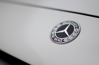 The image features a close-up of a Mercedes-Benz logo on a smooth, light-colored surface. The logo is in the shape of a three-pointed star within a circle, with the brand name inscribed around it.