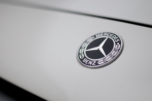 The image features a close-up of a Mercedes-Benz logo on a smooth, light-colored surface. The logo is in the shape of a three-pointed star within a circle, with the brand name inscribed around it.