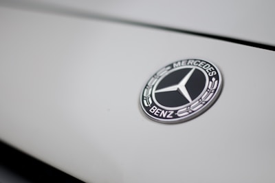 The image features a close-up of a Mercedes-Benz logo on a smooth, light-colored surface. The logo is in the shape of a three-pointed star within a circle, with the brand name inscribed around it.