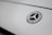 The image features a close-up of a Mercedes-Benz logo on a smooth, light-colored surface. The logo is in the shape of a three-pointed star within a circle, with the brand name inscribed around it.