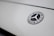 The image features a close-up of a Mercedes-Benz logo on a smooth, light-colored surface. The logo is in the shape of a three-pointed star within a circle, with the brand name inscribed around it.