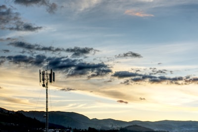 A telecommunications tower stands prominently against a backdrop of rolling hills and a dramatic sky filled with clouds. The sunlight casts a warm glow, creating a soothing yet dynamic contrast.