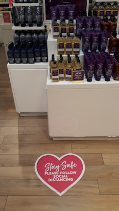 Shelves display a variety of personal care products in bottles and tubes, organized neatly in a store. A visible heart-shaped floor sign with a message encourages social distancing.