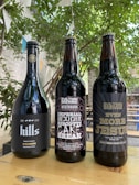 Three bottles of dark-colored beer placed on a wooden table outdoors. The bottle labels display creative designs and text, with a natural, leafy background and hanging decorative flags.