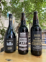 Three bottles of dark-colored beer placed on a wooden table outdoors. The bottle labels display creative designs and text, with a natural, leafy background and hanging decorative flags.