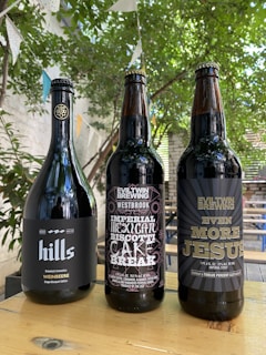 Three bottles of dark-colored beer placed on a wooden table outdoors. The bottle labels display creative designs and text, with a natural, leafy background and hanging decorative flags.