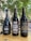 Three bottles of dark-colored beer placed on a wooden table outdoors. The bottle labels display creative designs and text, with a natural, leafy background and hanging decorative flags.