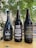 Three bottles of dark-colored beer placed on a wooden table outdoors. The bottle labels display creative designs and text, with a natural, leafy background and hanging decorative flags.