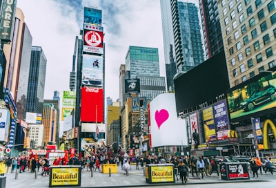 A bustling urban scene with towering skyscrapers and numerous digital billboards showcasing advertisements and entertainment content. A large crowd of people is visible, indicating a busy environment. Bright colors and diverse signage add to the vibrant atmosphere.