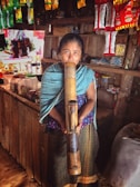 A woman stands inside a small shop holding a large bamboo pipe. The shop is adorned with hanging packets of snacks and a variety of goods on the shelves. The woman wears traditional clothing with a patterned blue shawl and skirt.