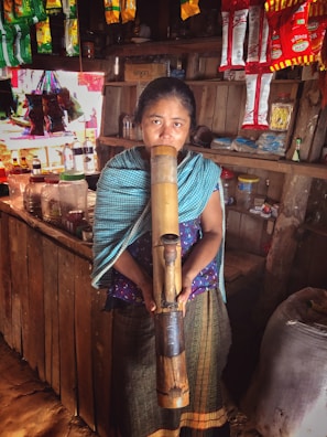 A woman stands inside a small shop holding a large bamboo pipe. The shop is adorned with hanging packets of snacks and a variety of goods on the shelves. The woman wears traditional clothing with a patterned blue shawl and skirt.