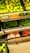 Fresh produce is neatly arranged in black crates at a market or grocery store. The top crate displays a large quantity of lemons or limes. Below them, there are crates filled with green chilies, garlic, and shallots or small onions. Each crate has a pricing label in a rectangular holder.