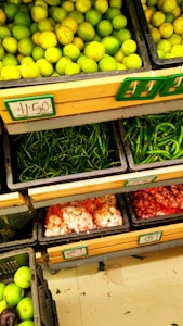 Fresh produce is neatly arranged in black crates at a market or grocery store. The top crate displays a large quantity of lemons or limes. Below them, there are crates filled with green chilies, garlic, and shallots or small onions. Each crate has a pricing label in a rectangular holder.