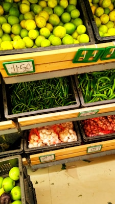 Fresh produce is neatly arranged in black crates at a market or grocery store. The top crate displays a large quantity of lemons or limes. Below them, there are crates filled with green chilies, garlic, and shallots or small onions. Each crate has a pricing label in a rectangular holder.