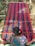 A person is weaving a colorful textile on a traditional loom, with vibrant stripes of red, blue, and green. The textile is stretched across a wooden frame, and the person is wearing a patterned blue garment.