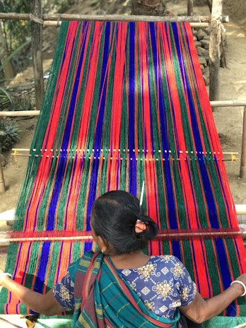 A person is weaving a colorful textile on a traditional loom, with vibrant stripes of red, blue, and green. The textile is stretched across a wooden frame, and the person is wearing a patterned blue garment.
