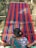 A person is weaving a colorful textile on a traditional loom, with vibrant stripes of red, blue, and green. The textile is stretched across a wooden frame, and the person is wearing a patterned blue garment.