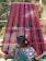 A person is weaving a colorful textile on a traditional loom, with vibrant stripes of red, blue, and green. The textile is stretched across a wooden frame, and the person is wearing a patterned blue garment.