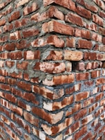 A close-up view of a brick wall corner, with roughly textured red bricks and visible gray mortar between them, creating a rugged and sturdy appearance.