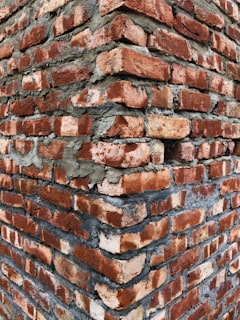 A close-up view of a brick wall corner, with roughly textured red bricks and visible gray mortar between them, creating a rugged and sturdy appearance.