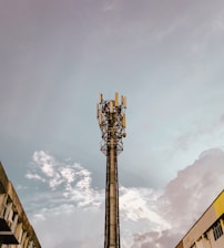 A tall communication tower stands prominently against a backdrop of a cloudy sky. The tower is equipped with multiple antennas and electronic equipment, suggesting its function in telecommunications. Surrounding the tower, there are parts of buildings visible in the lower corners of the image, indicating an urban setting.